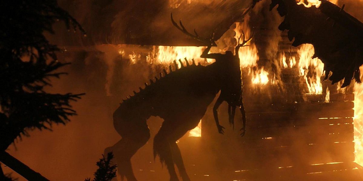 A elk-like monster composed of human body parts hunches in front of a burning cabin