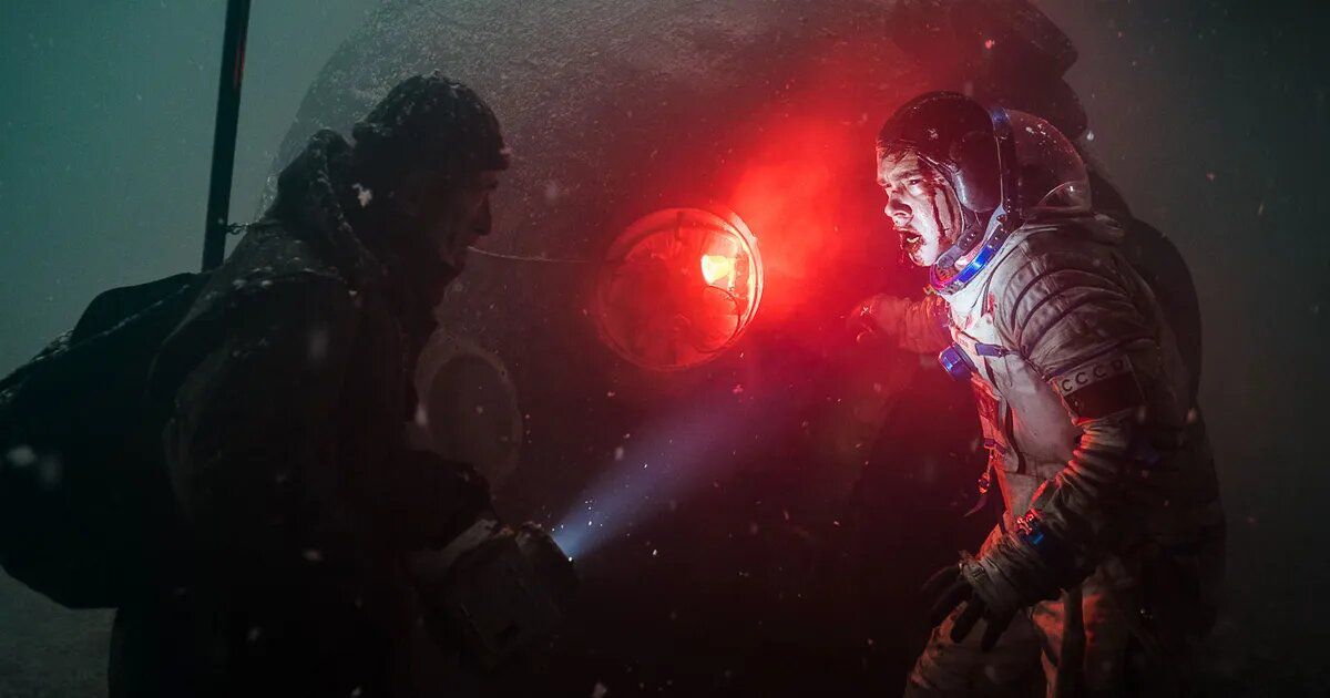 A helmetless man in a bloodied astronaut suit scowls at a man with a flashlight in front of a downed space capsule with an eerie red light emanating from its porthole.
