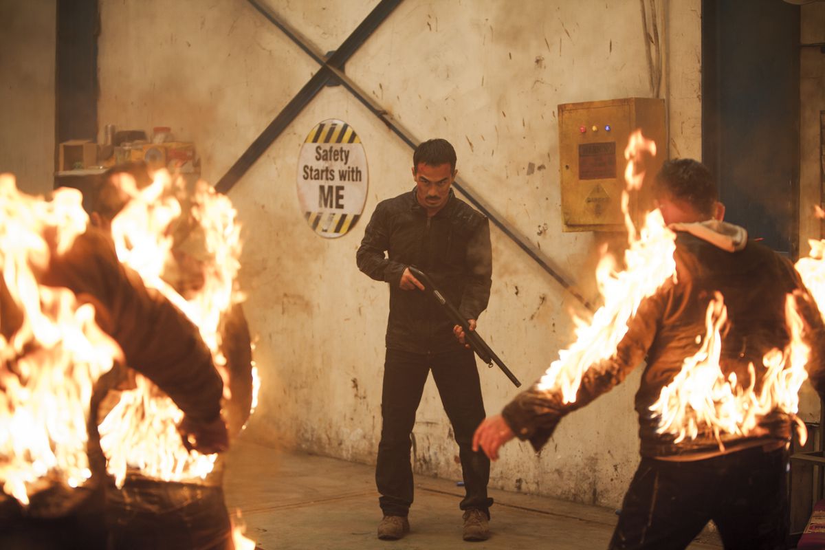 Joe Taslim stands in front of a “Safety starts with me” sign toting a shotgun facing several men on fire in The Night Comes for Us.