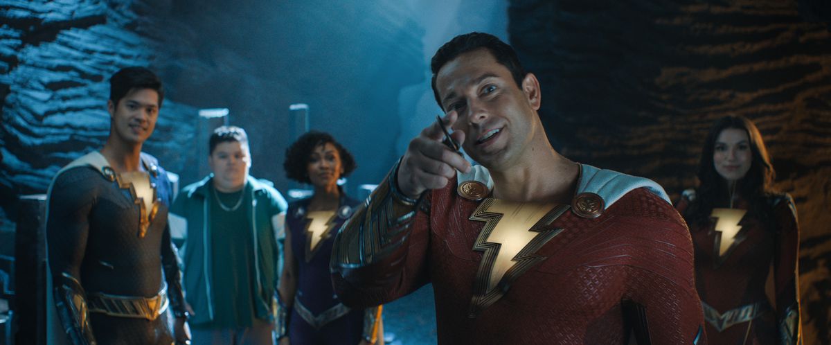 Zachary Levi när Shazam pekar in i kameran och ler, med medlemmar av sin hjältefamilj bakom sig, i Shazam!  Gudarnas raseri