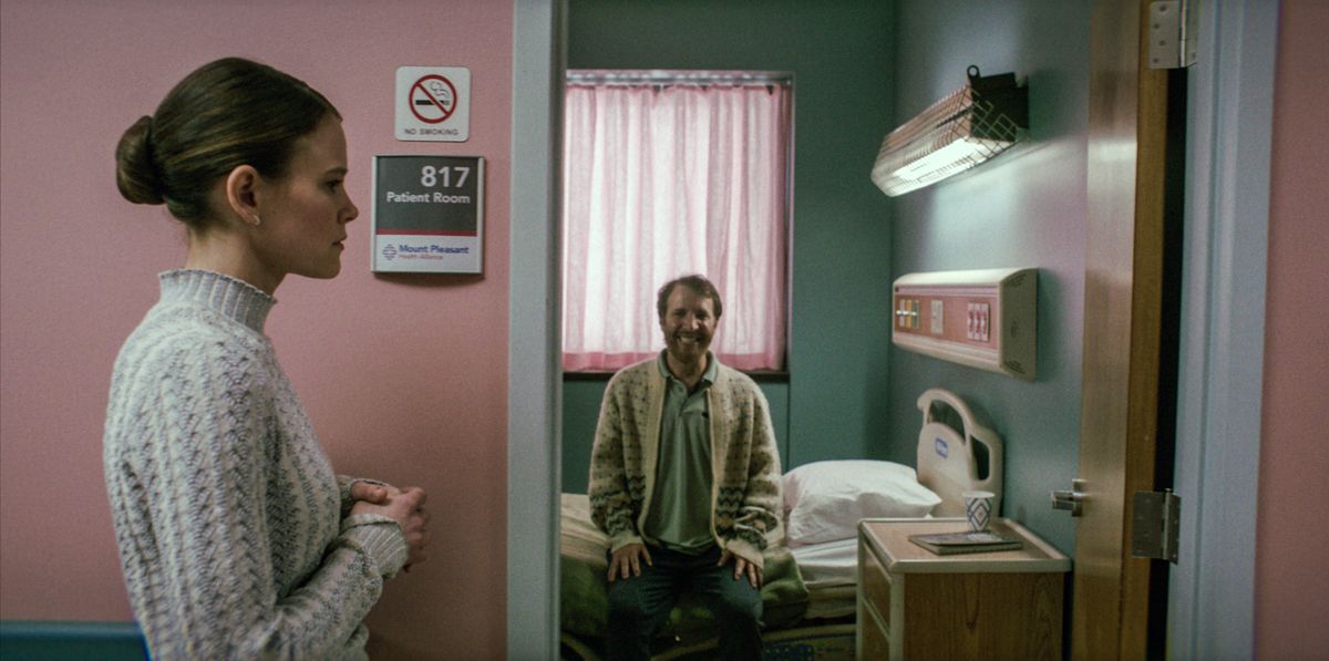 Sosie Bacon as Rose standing outside a hospital room in Smile. The man inside the room is sitting upright on the edge of the bed and smiling in an unsettling way.