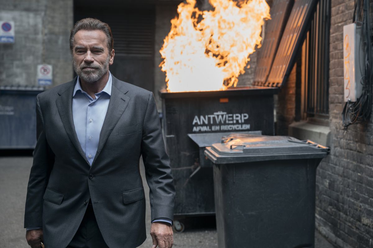 Arnold Schwarzenegger, wearing a grey suit and blue shirt, walks away from a literal dumpster fire in the Netflix series FUBAR