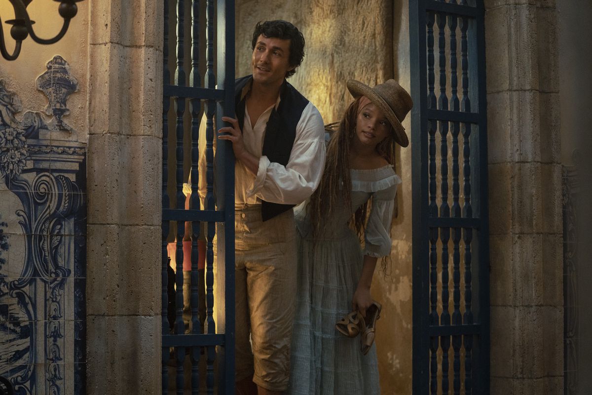 Jonah Hauer-King and Halle Bailey peer out from behind a gate in The Little Mermaid. Halle Bailey wears a straw hat and a light blue dress, while Hauer-King is in his classic Prince Eric attire.