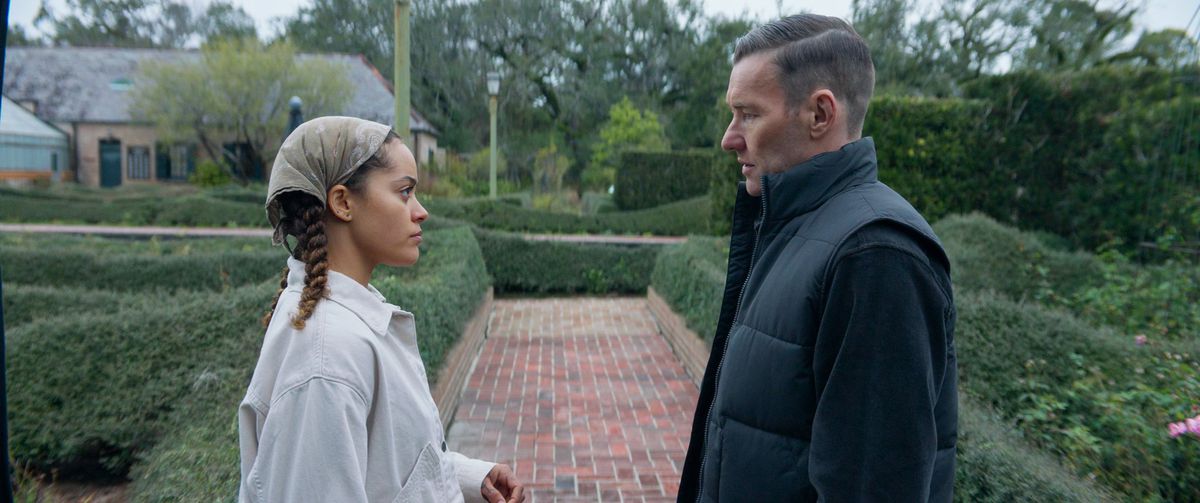 Quintessa Swindell and Joel Edgerton star at each other on a brick pathway, surrounded by a green garden, in Master Gardener.