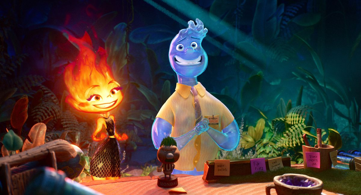 In an image from Pixar’s Elemental, Ember the fire woman and Wade the water guy smile. Wade looks like he’s pleading, with his hands put together and a white collared shirt on, while Ember smiles while wearing a black dress. Plants are behind them, while a wooden desk is in front of them.