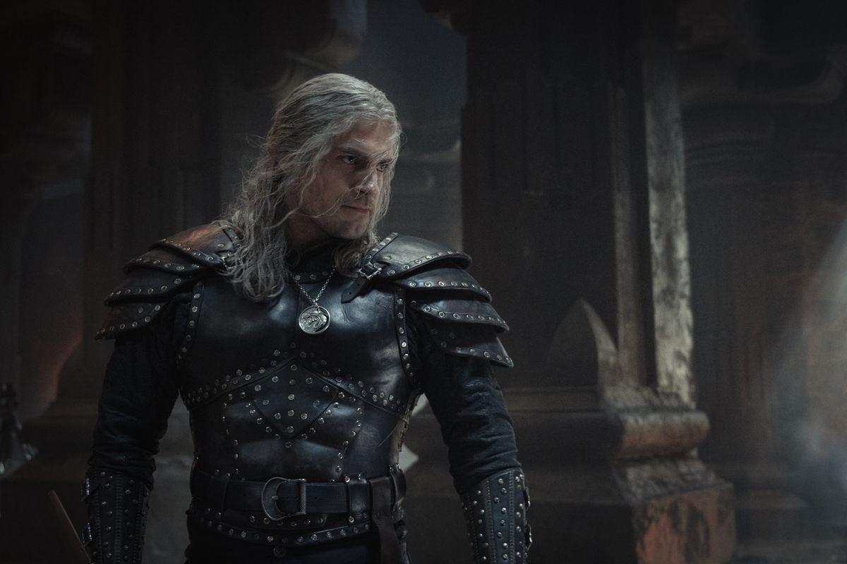 Henry Cavill poses in big black studded armor in a castle type area looking ready to punch a guy in The Witcher