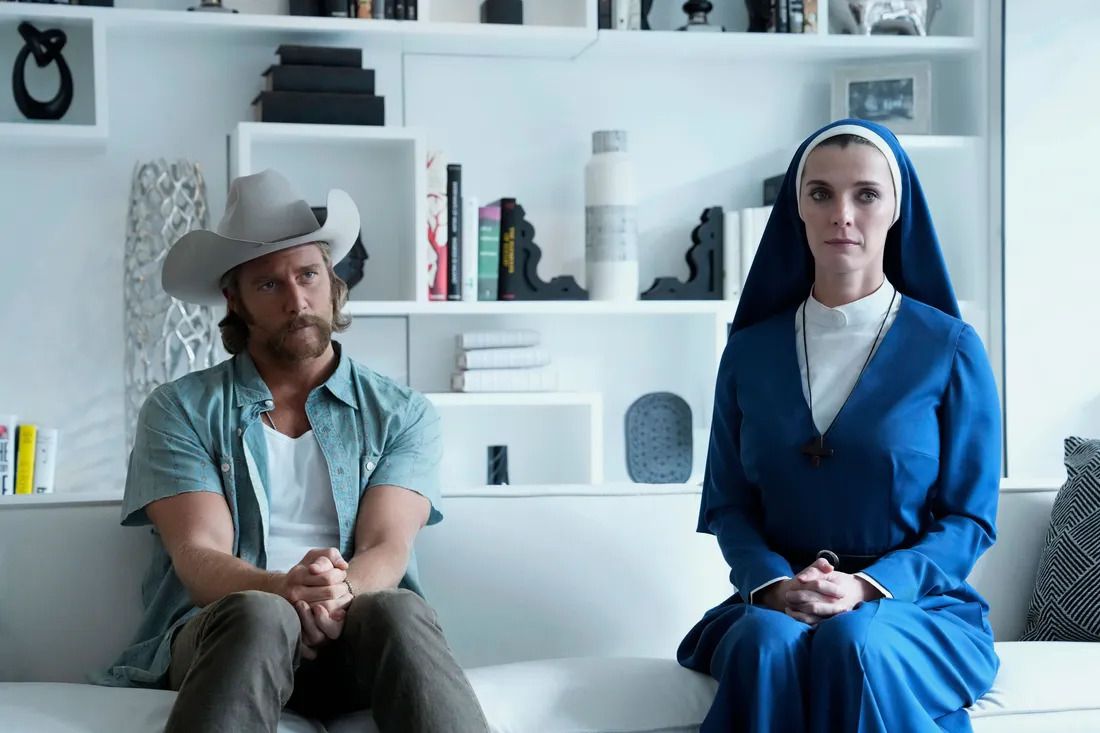 (L-R) A man in a cowboy hat (Jake McDorman) and a woman in a blue nun habit (Betty Giplin) sit on a white couch in a modernist living room in Mrs. Davis.