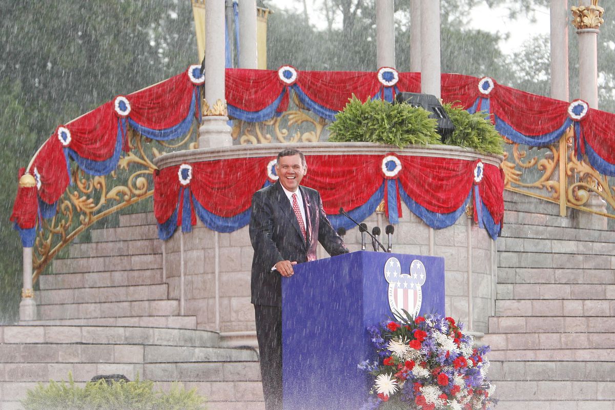 Den amerikanska senatorn Mel Martinez står och ler i hällande regn vid en utomhuspodium med ett Musse Pigg-huvud med den amerikanska flaggan på vid en massnaturaliseringsceremoni i Walt Disney World i Florida den 4 juli 2007