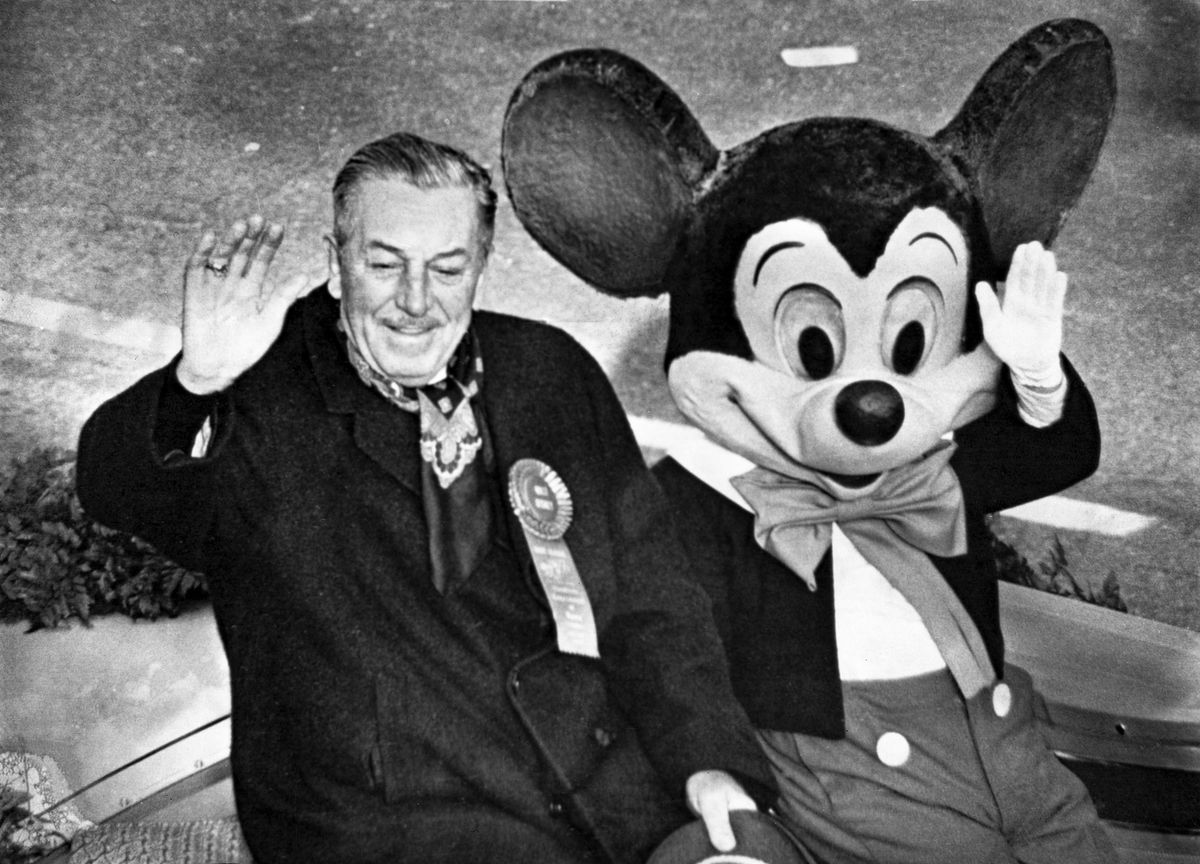 En bild på Walt Disney som sitter bredvid en utklädd artist som Musse Pigg 1966 som Grand Marshal of the Rose Parade, med båda vinkar mot publiken