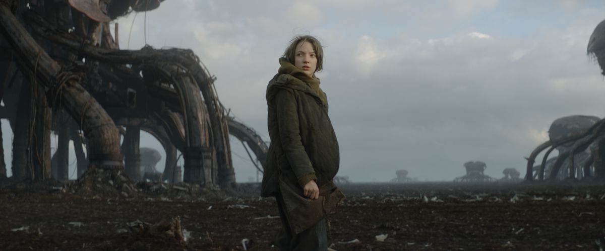 13-year-old Vesper (Raffiella Chapman) stands in a barren field with giant rusty octopus robots on the horizon in Vesper