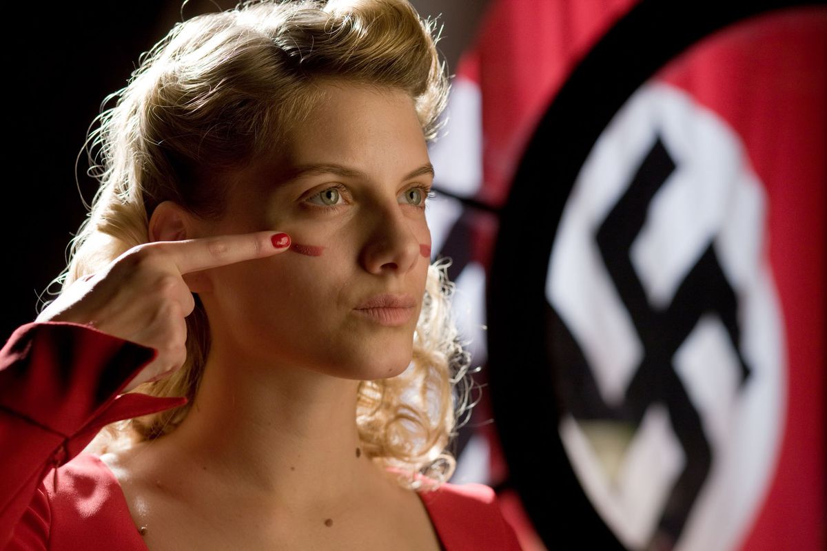 A young blond woman in a red dress smears warpaint on her face with a Nazi Swastika hanging in the background