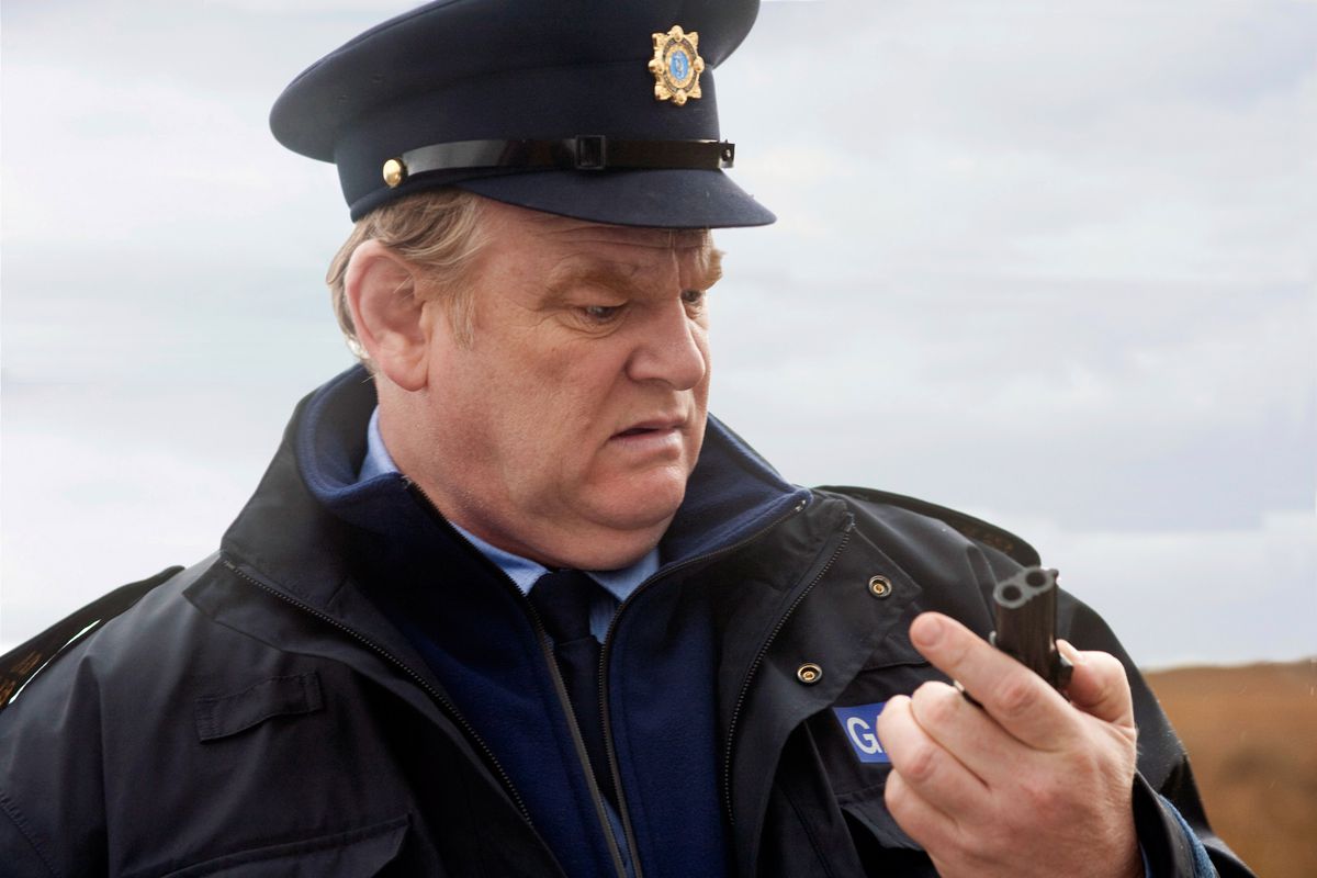 Brendan Gleeson, dressed as a policeman, looks in consternation at a small gun in his hand