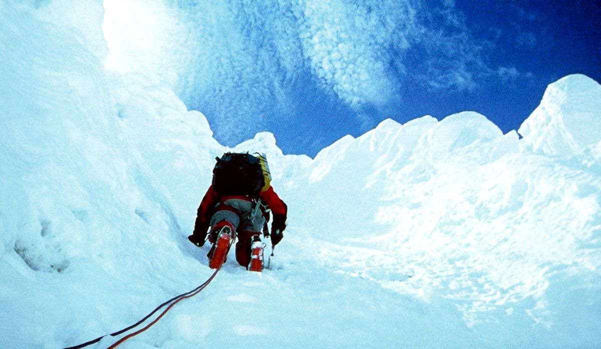A mountain climber, shot from below, scales a sheer ice face, with blue sky and clouds above