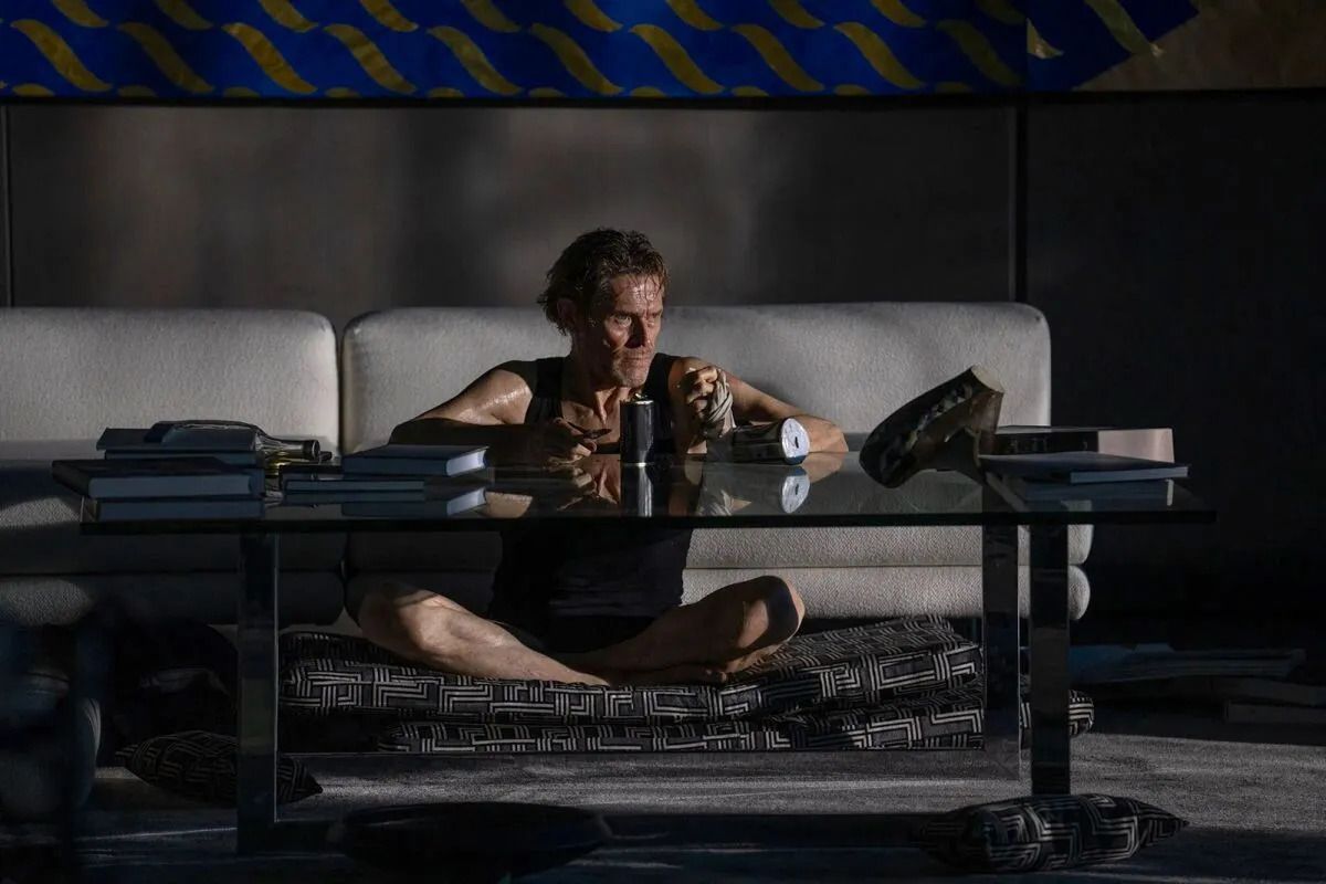En naken, rufsig man i boxerkalsonger (Willem Dafoe) sitter med korslagda ben framför ett glasbord i ett mörkt vardagsrum med ljus som strömmar in från ett fönster utanför skärmen i Inside.