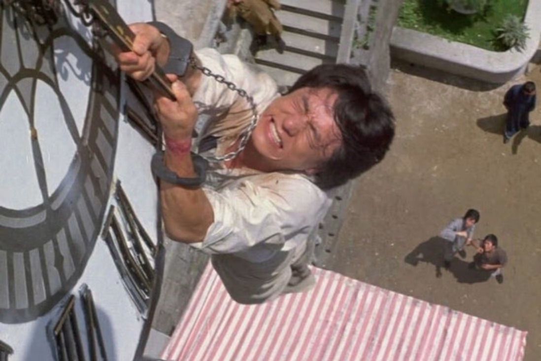 A man (Jackie Chan) visibly strains to hold on to a chain dangling from the face of a clocktower with two men looking on in astonishment and concern from ground-level.