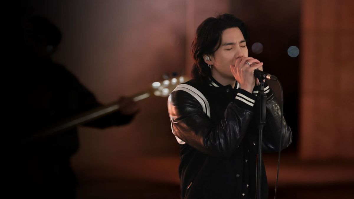 Suga från BTS sjunger med slutna ögon och båda händerna virade runt en mikrofon medan han bär en svartvit jacka i Suga: Road to D-Day.