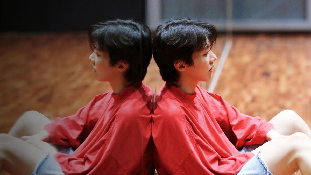 BTS-rapparen J-Hope lutar sig mot en golvlång spegel iförd jeansshorts och en ljusröd långärmad skjorta.  Vi ser hans spegelbild i spegeln.