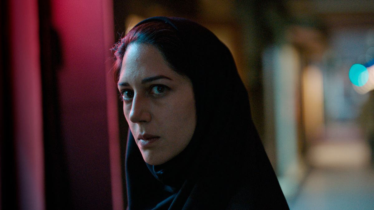 Arezoo Rahimi (Zahra Amir Ebrahimi) står mitt i en mörk korridor med ljus som reflekteras på sidan av hennes ansikte, hennes mörka hår delvis skymt av en huvudduk i Holy Spider.