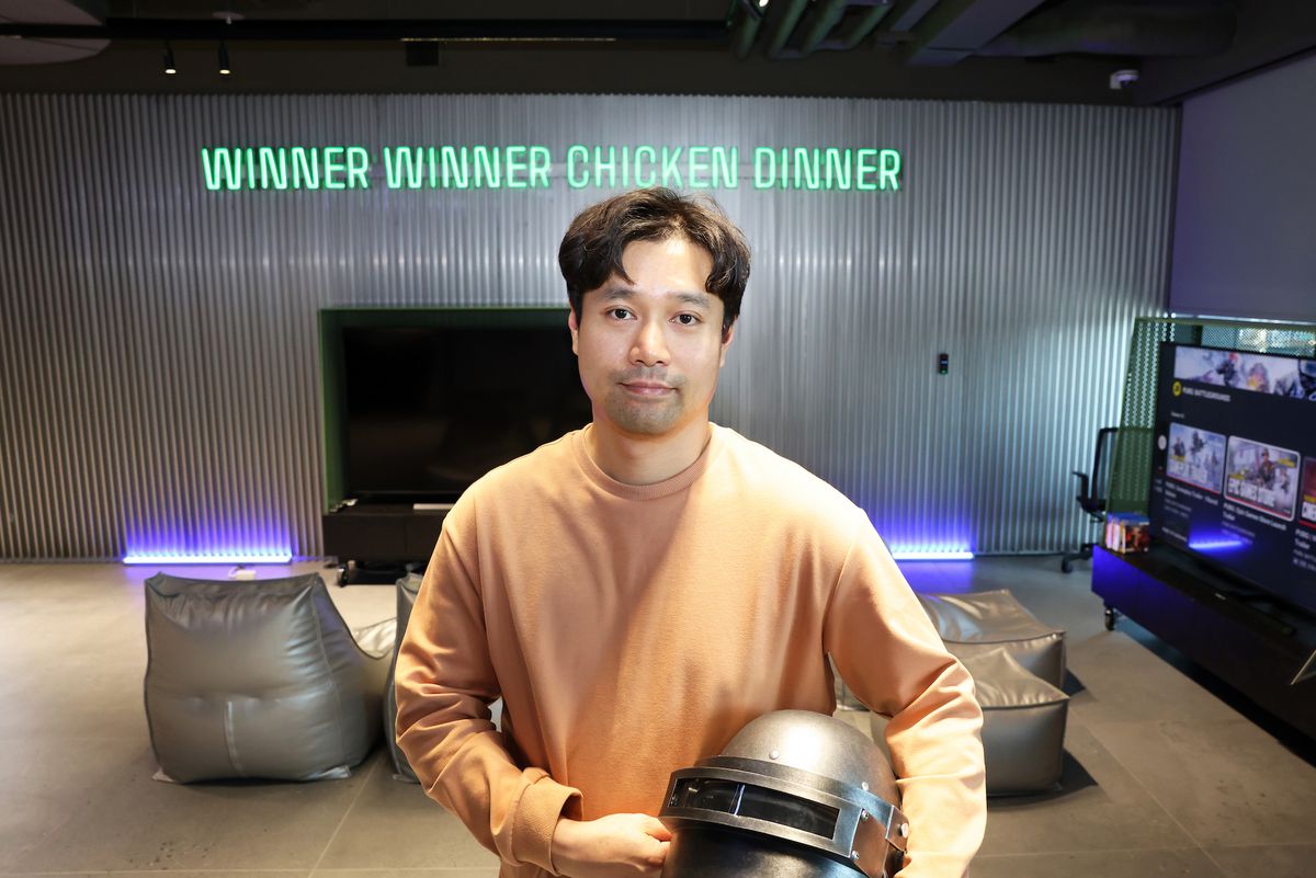 Ett fotografi av Taeyhun Kim från PUBG Studios som håller den klassiska PUBG-hjälmen och står framför en neonskylt där det står 