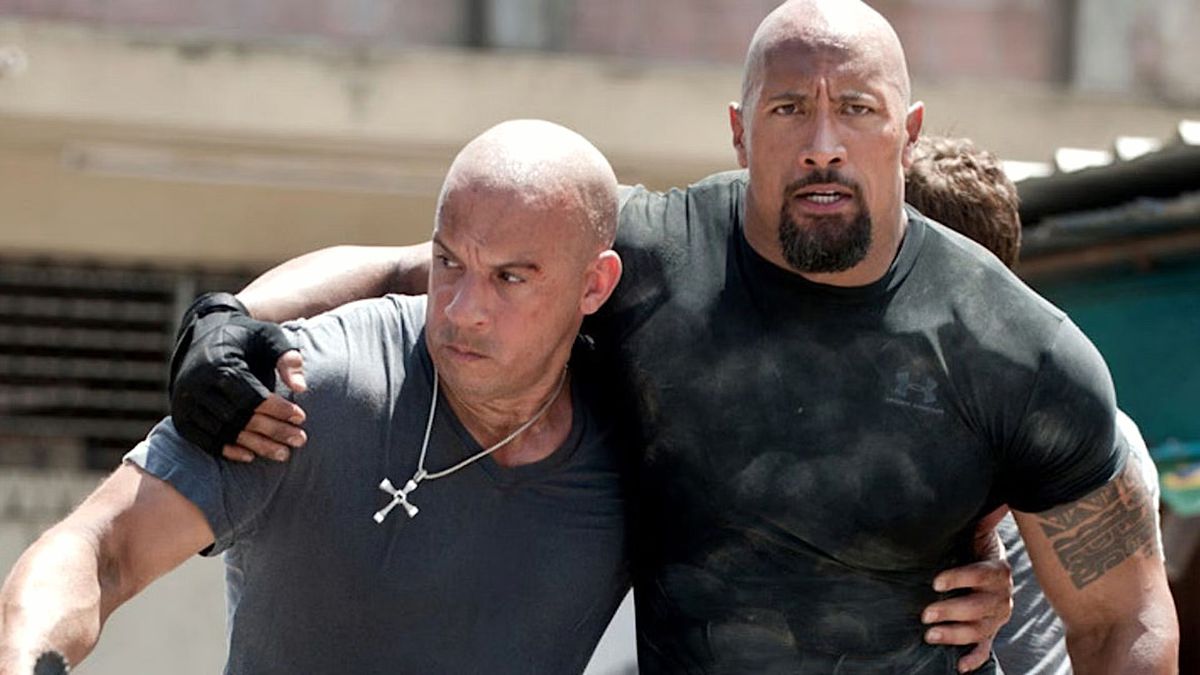 Dom (Vin Diesel) och Hobbs (Dwayne Johnson) stödjer varandra med armarna runt varandras midjor i Fast Five.  Doms krucifix, som för närvarande inte lämnats ut till en medlem av hans familj för förvaring, hänger framträdande runt hans hals.