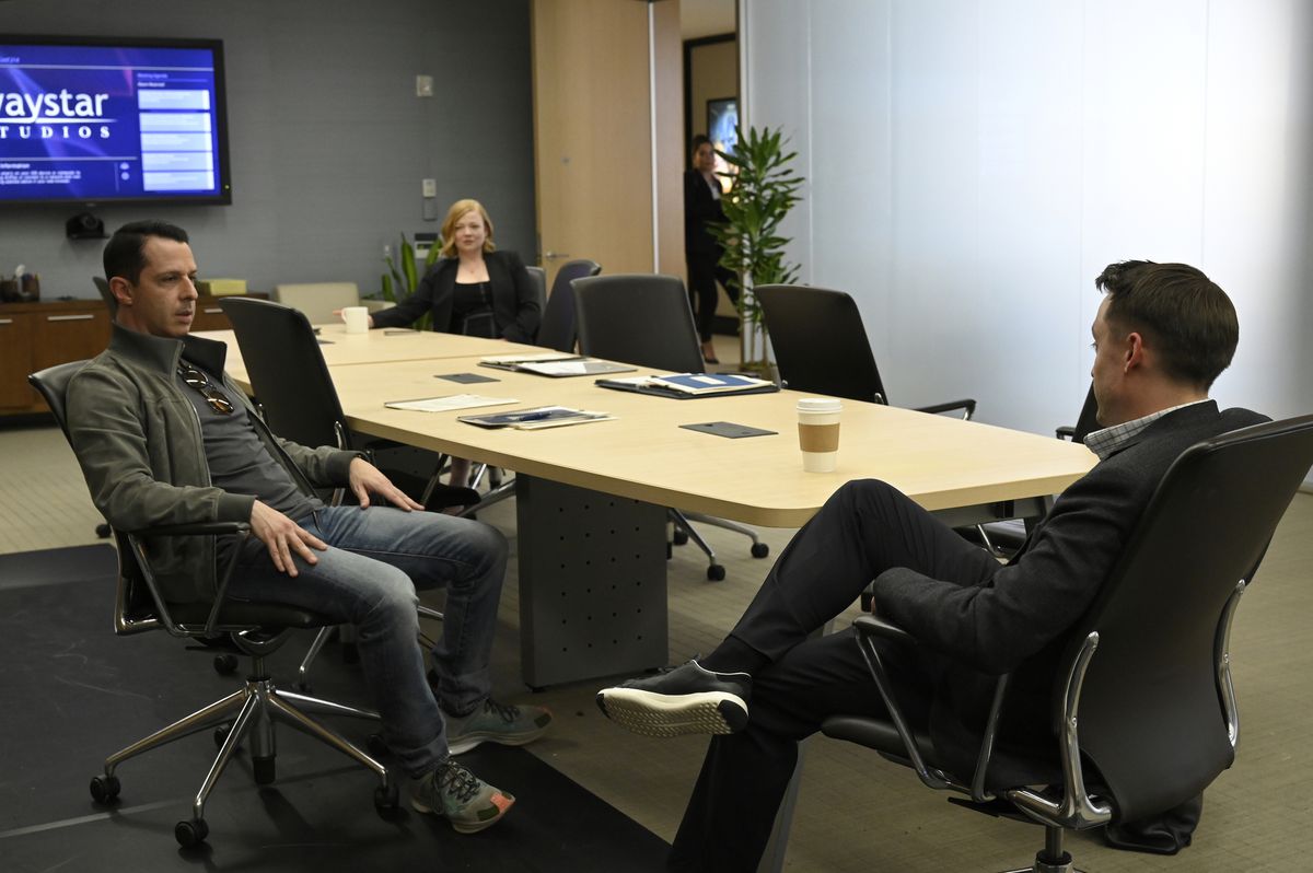 Shiv (Sarah Snook) sitter på andra sidan i bakgrunden vid ett konferensbord medan hennes bröder Kendall (Jeremy Strong) och Roman (Kieran Culkin) sitter och pratar närmare förgrunden