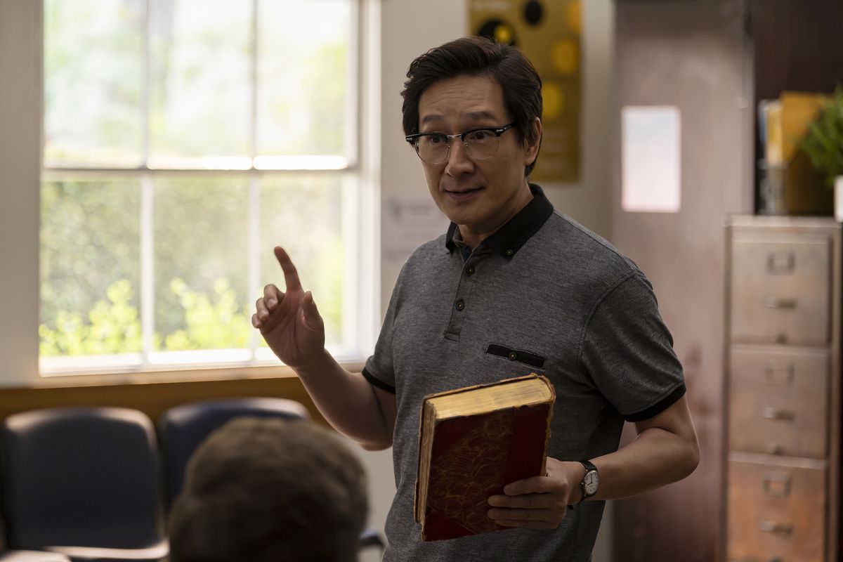 Ke Huy Quan på amerikansk född kinesiska.  Han håller en bok i handen och verkar undervisa framför en klass.