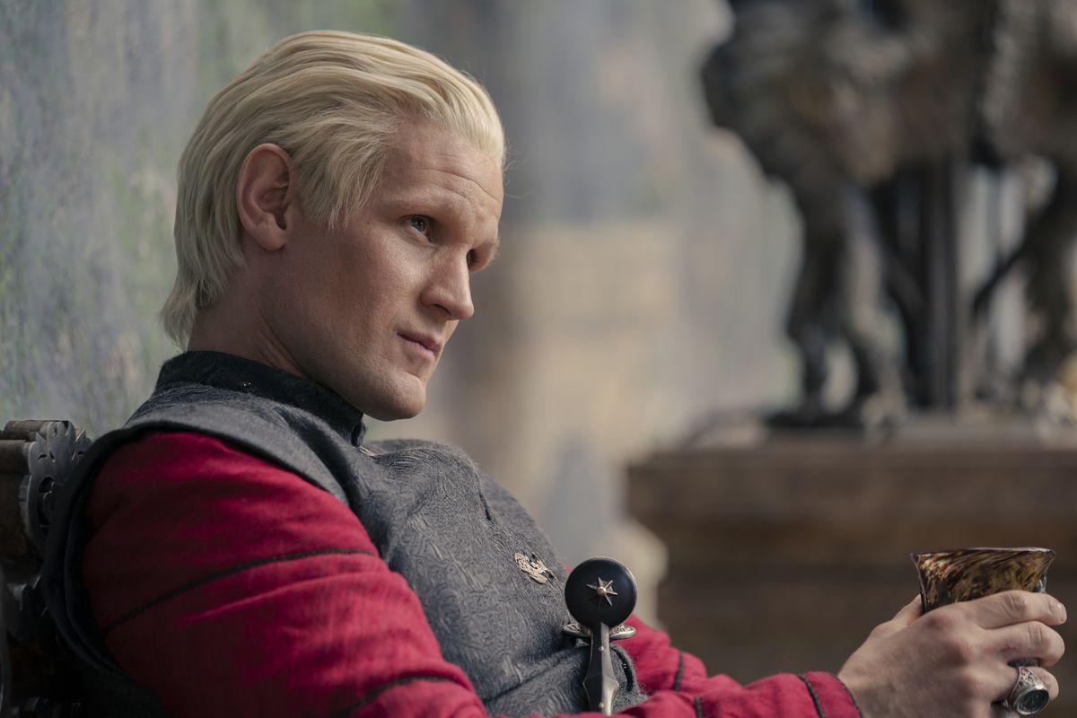 Daemon Targaryen, smord, iklädd röd skjorta och grå mantel, stirrar på någon när han greppar sitt vin i House of the Dragon