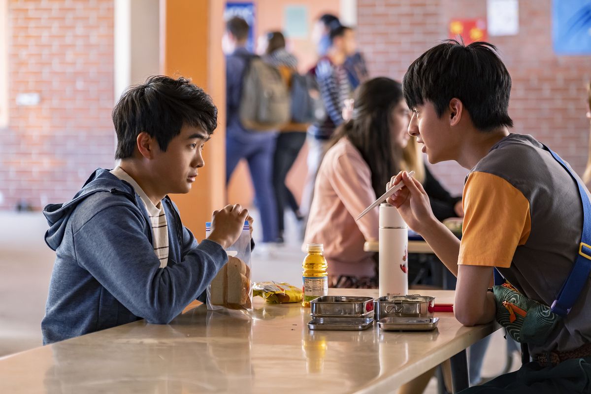 Ben Wang och Jim Liu som Jin Wang och Wei-Chen sitter mitt emot varandra vid ett skollunchbord.  Jin har en smörgås med vitt bröd och äppeljuice, medan Wei-Chen har en metallburk med mat och äter med pinnar. 