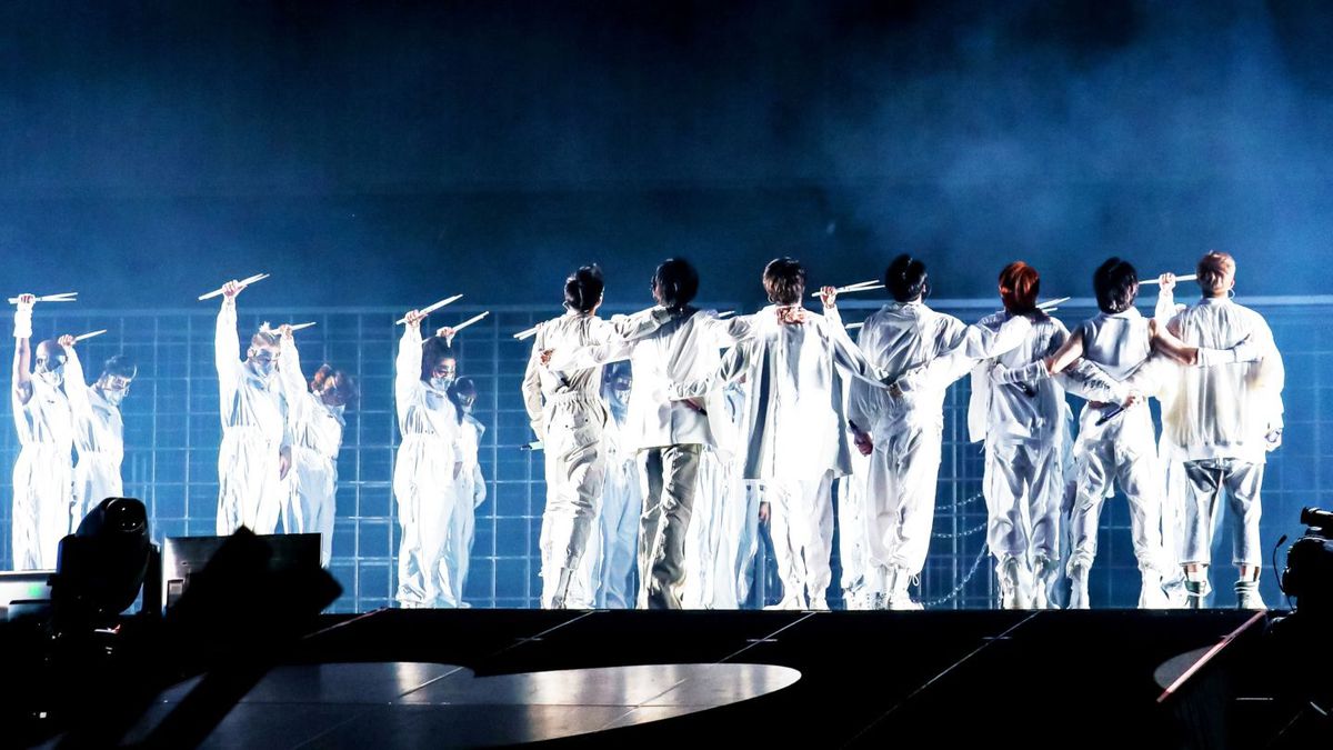 Medlemmarna i BTS bär helvitt, med armarna runt varandra, i en vy bakifrån på deras Permission to Dance on Stage LA-konsert.  Bakgrundsdansare står med armarna i vädret.  Alla håller i trumpinnar.