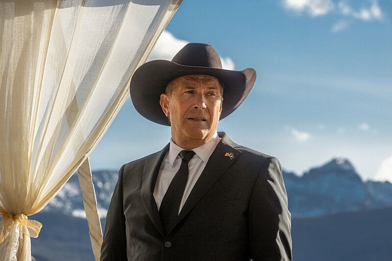 Kevin Costner i Yellowstone, stående vänd mot kameran iförd svart kostym med svart cowboyhatt