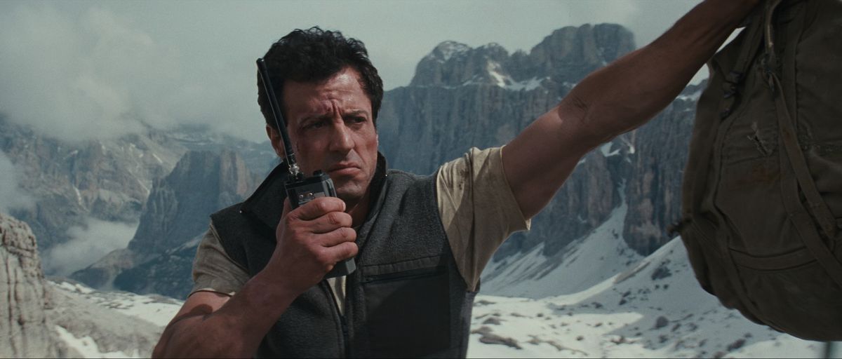Sylvester Stallone talks on a radio while resting his arm on a cliff side in Cliffhanger. Behind him is a gorgeous view of snowy mountains.