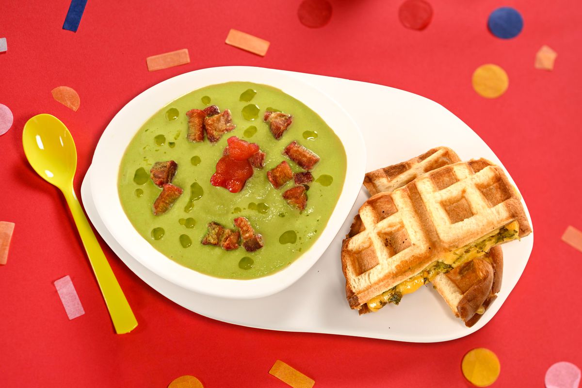 En skål med grön soppa med flytande bitar.  Bredvid står en våffelmacka. 