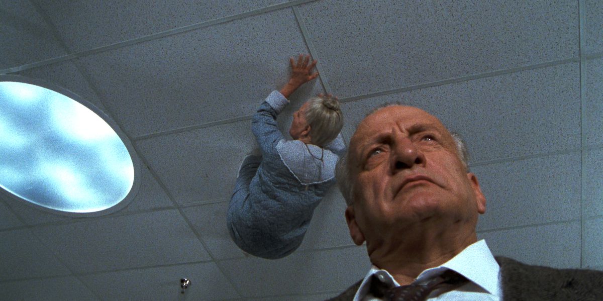 George C. Scott walks around a hospital, oblivious, as a gray-haired woman crawls across the ceiling above him in Exorcist III