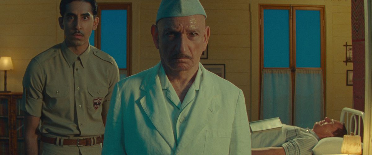 Dev Patel, dressed in the taupe uniform of a British military officer in occupation-era India, stands behind Ben Kingsley in blue scrubs and cap as an Indian doctor, as Benedict Cumberbatch lies in bed, flat on his back, behind them both in Netflix’s short Poison