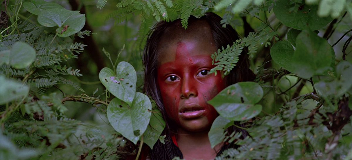 Ett barn klädt i röd ansiktsfärg tittar genom ett tak av löv i Baraka.