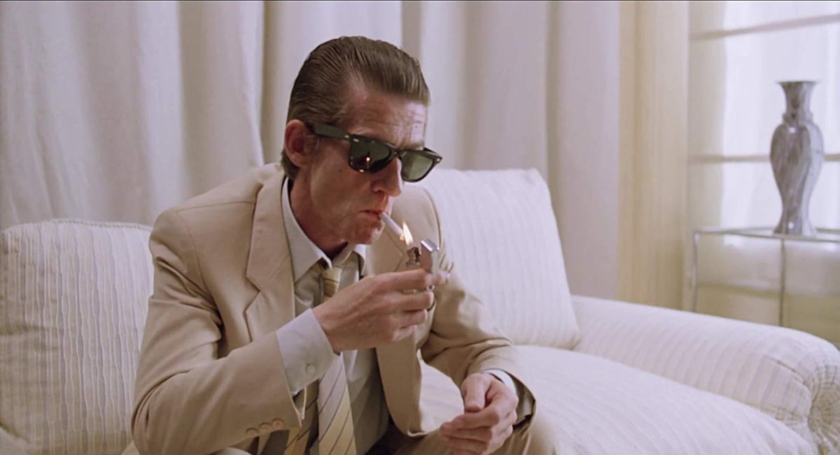 John Hurt som Braddock tänder en cigarett och sitter på en vit soffa med solglasögon på i The Hit.