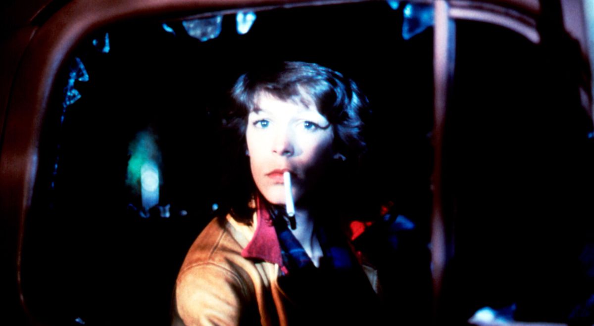 Jamie Lee Curtis, cigarette dangling from her lips, sits calmly in a car with the window smashed out in John Carpenter’s The Fog