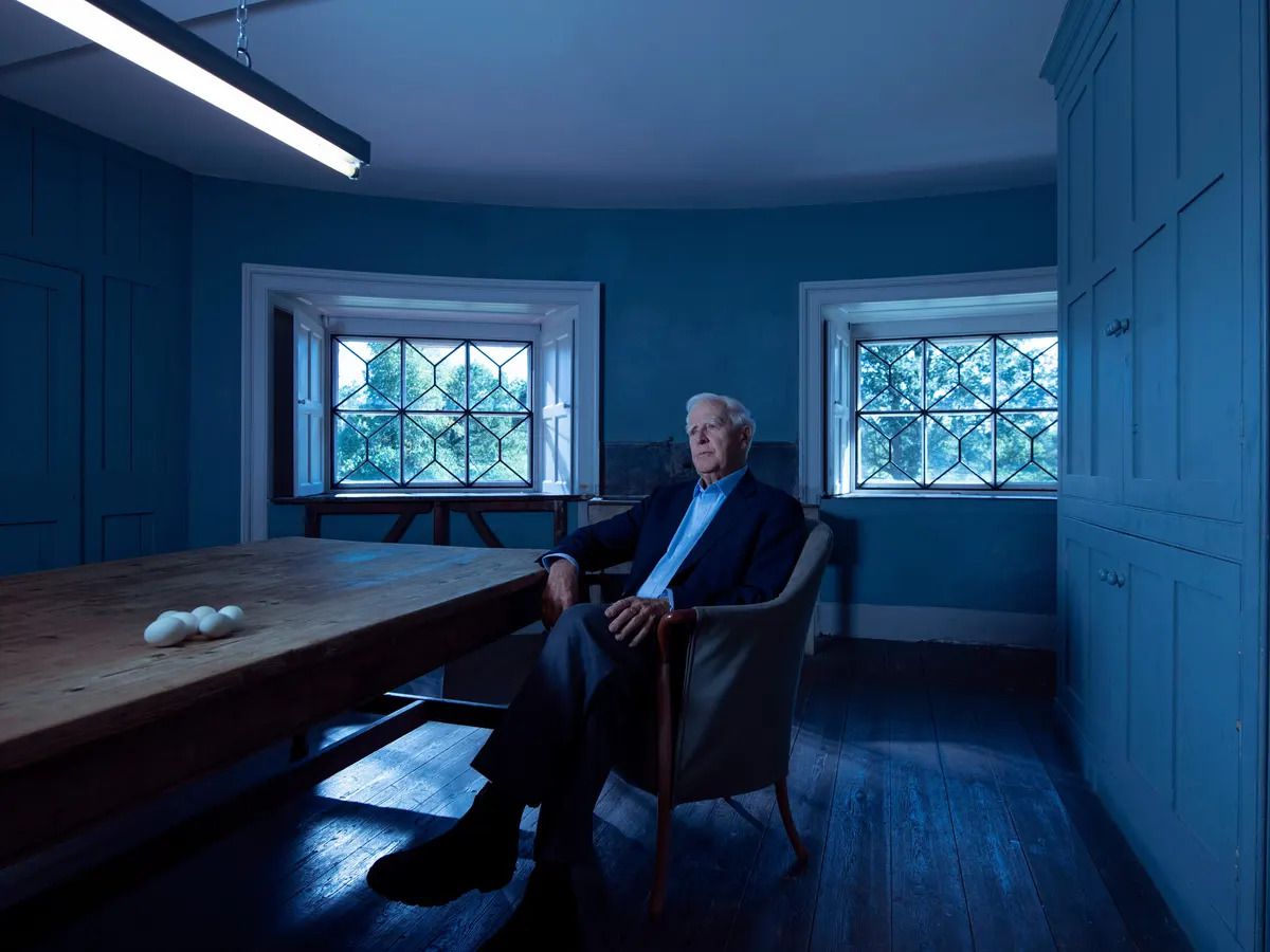 John le Carré sitting in a chair next to a set of eggs on a wooden table in The Pigeon Tunnel.