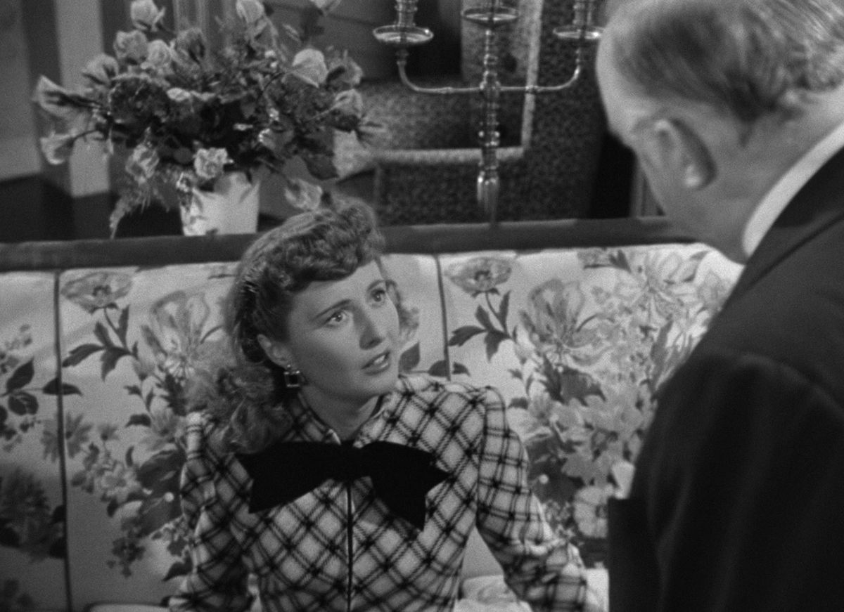 Barabara Stanwyck sits on a couch in Christmas in Connecticut. She wears a long sleeve top with a large bow tie and looks in confusion at a man standing above her.