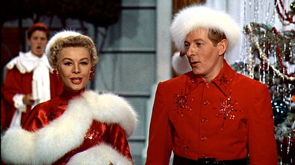 Vera-Ellen as Judy and Danny Kaye as Phil in White Christmas, dressed in festive red outfits adorned with sparkly red snowflakes and white fur, standing in front of a Christmas tree decorate d with tinsel.