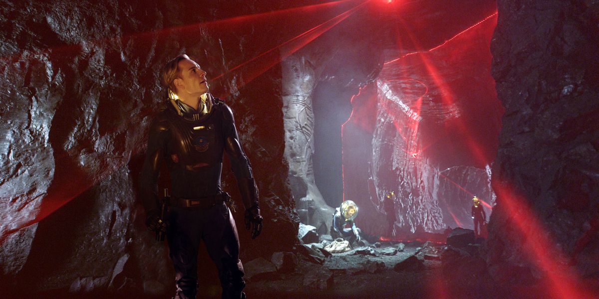 Michael Fassbender, in a futuristic full-body black bodysuit, stands in a black stone cavern and looks up at a series of red lasers mapping the space as other people in spacesuits wander behind him in Prometheus 
