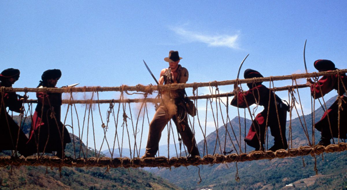 Indiana Jones (Harrison Ford), in the usual battered fedora and holding a sword, stands in the middle of a rope bridge with turbaned men in red approaching him from either side with swords and guns in Indiana Jones and the Temple of Doom