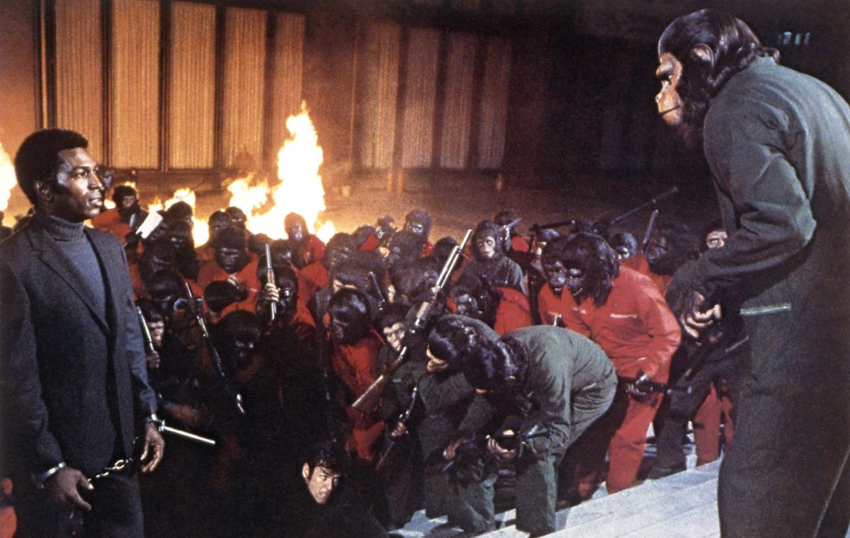Caesar the ape (Roddy McDowall) stands in front of a crowd of other chimps and apes wearing jumpsuits, carrying rifles, and beating a man to the ground, as flames burn behind them. Caesar is making eye contact with McDonald (Hari Rhodes), a Black man in a suit and handcuffs at the front of the crowd. From Conquest of the Planet of the Apes.