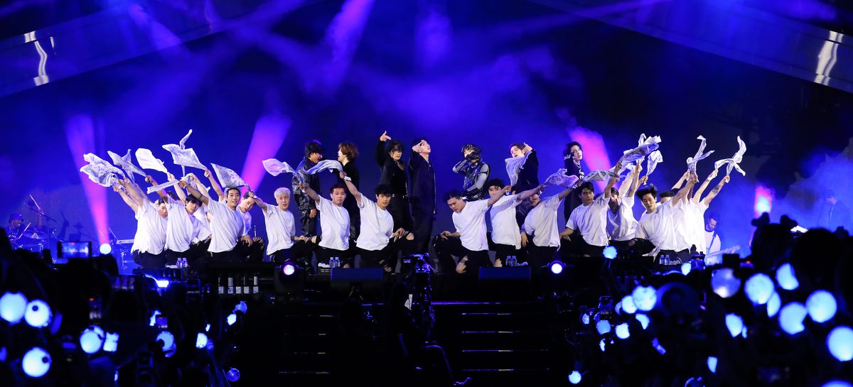 Medlemmarna i BTS, på scenen under Yet to Come-konserten, bakom en rad knästående manliga dansare i svarta byxor och vita t-shirts, viftande med vitt tyg