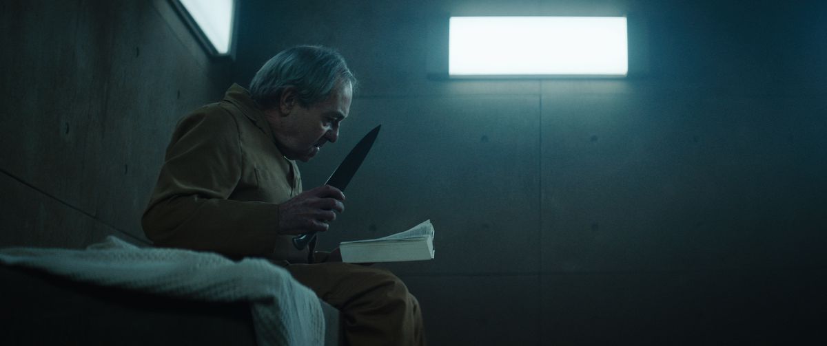 En äldre man som håller fast en otrevlig kniv och läser en bok sitter på sin säng i en svagt upplyst betongcell i plattformen.