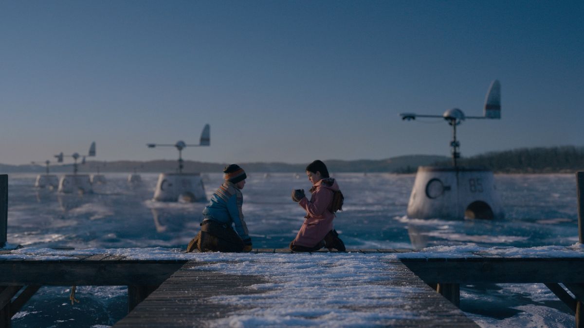 Två barn knä i slutet av en brygga vid en frusen sjö full av mystiska numrerade anordningar som ser ut som de snurrande tornen i vattna ubåtar.