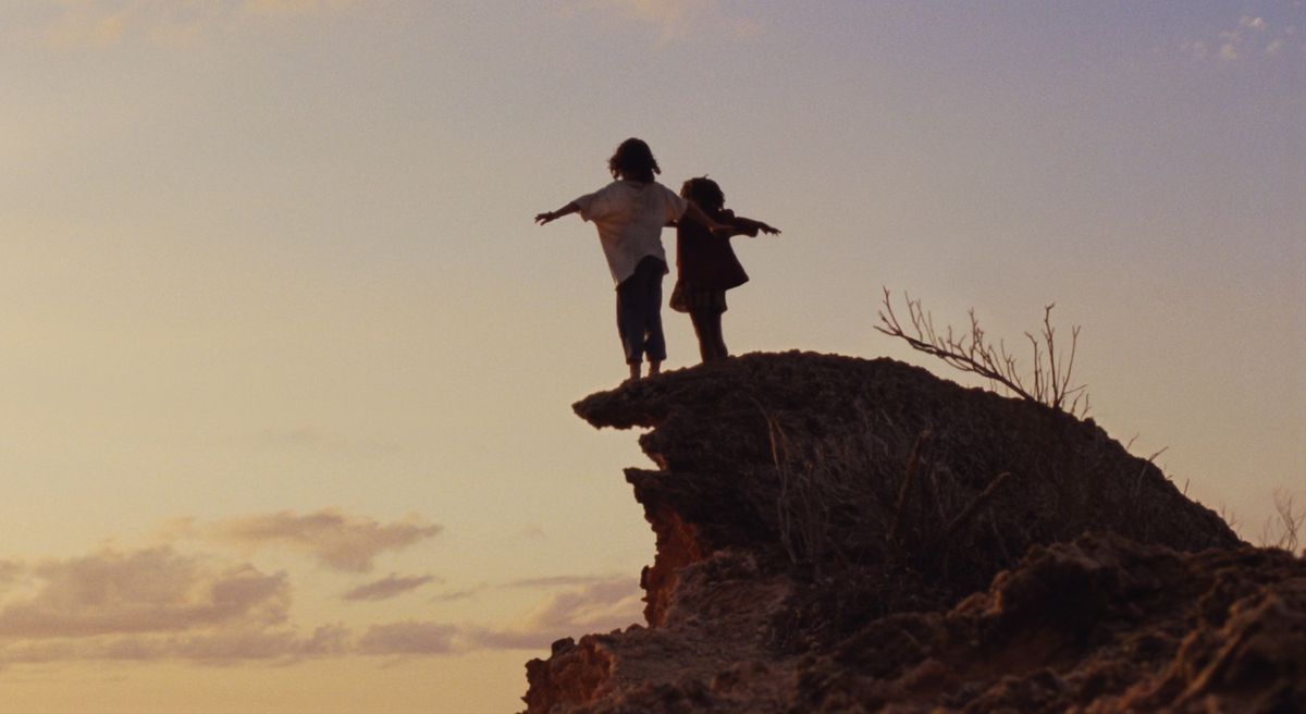 En flicka och en pojke står på en karg, rödaktig klippa vid solnedgången, vapnar ut som om de tänker flyga, silhuett mot en mestadels tom himmel.