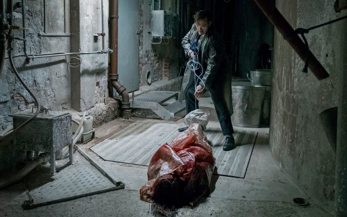 Matt Fillon, i kostym och regnrock av plast, drar ett plastförpackat, blodblött kropp genom ett smutsigt källarutrymme i huset som Jack byggde.