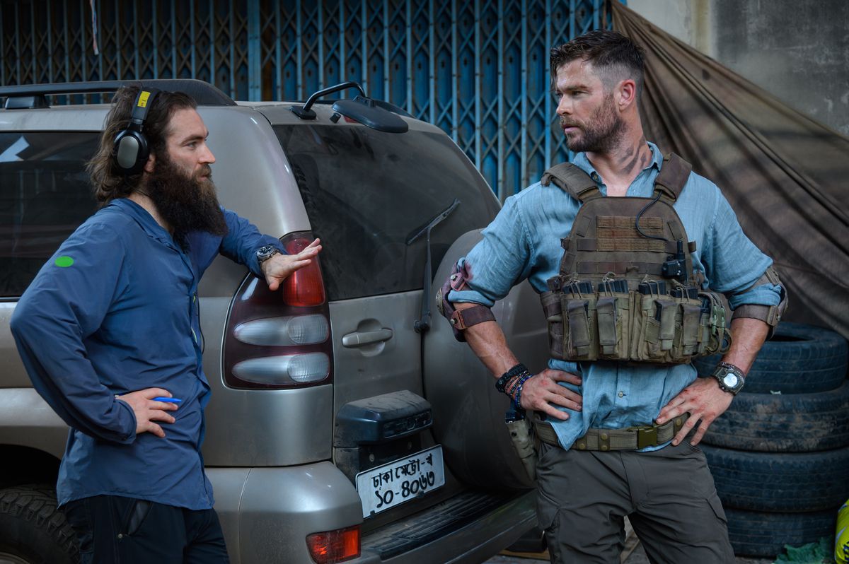 Sam Hargrave, som har ett gigantiskt skägg, lutar sig på en bil för att prata med sin knopp Chris Hemsworth, som har händerna på höfterna