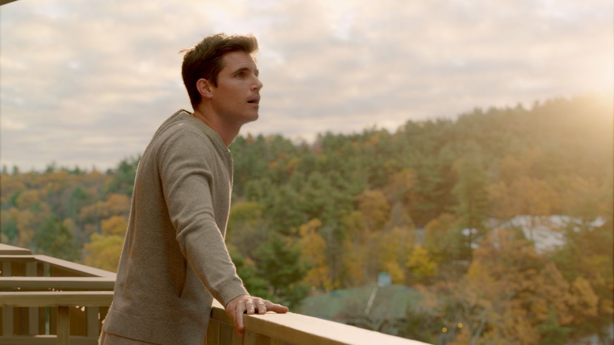 Robbie Amell står på en balkong med munnen hängande öppen och ser ut på en frodig skog som lyser med solljus.